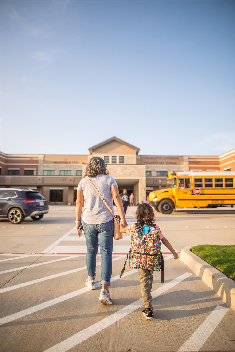  parent and child holding hands walking into school, school bus in the front of the school 